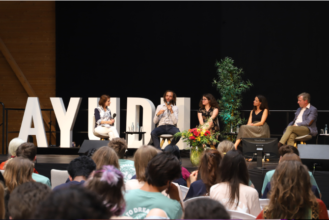A panel of speakers onstage in front of the AYUDH logo