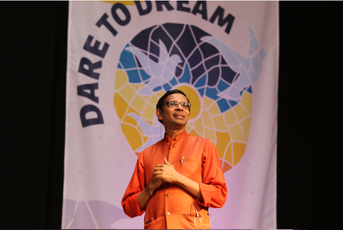 Swami Shubamritananda Puri addresses the crowd, standing in front of the AYUDH Dare to Dream backdrop. 