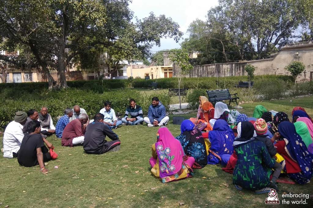 A large group of village men and women sit on the grass in discussion