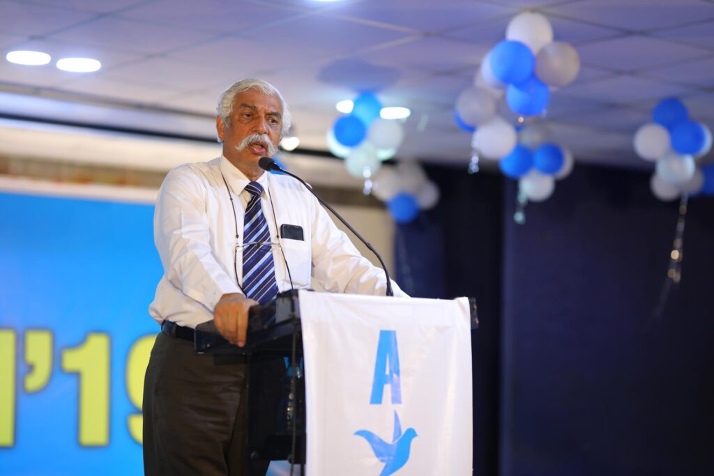 Speaker in slacks and tie stands at podium with blue and white balloons in the background. 