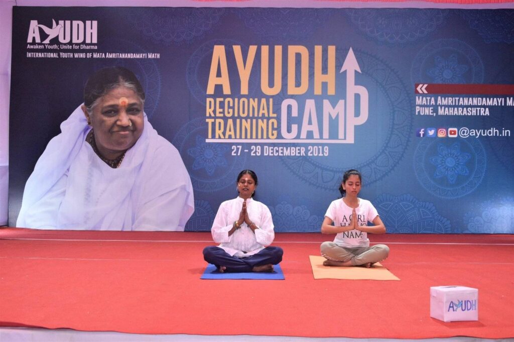 Two women sitting cross-legged on stage with their palms together.