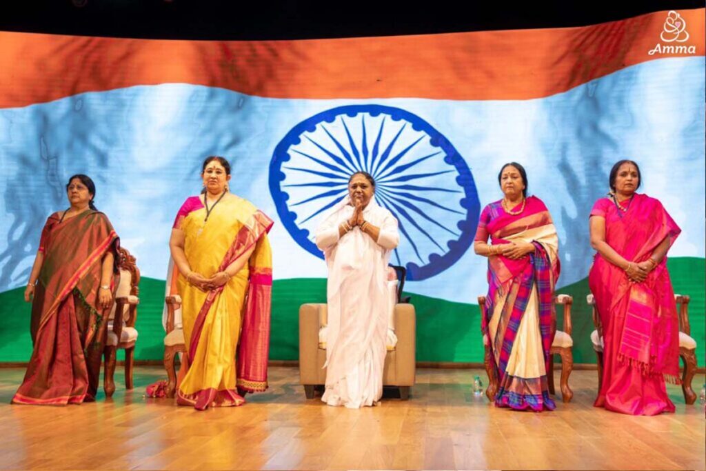 Amma stands in front of Indian flag with 4 other women, hands held in prayer