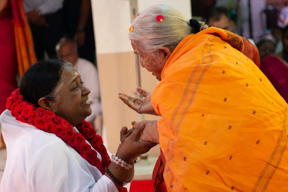 A woman in an orange sarti stands in front of Amma as she holds Amma's hand