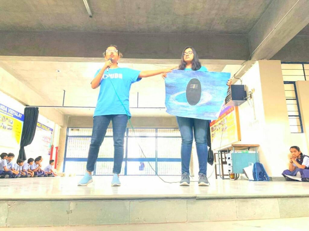 Two AYUDH women present with the Shivalinga poster on a stage to the students.