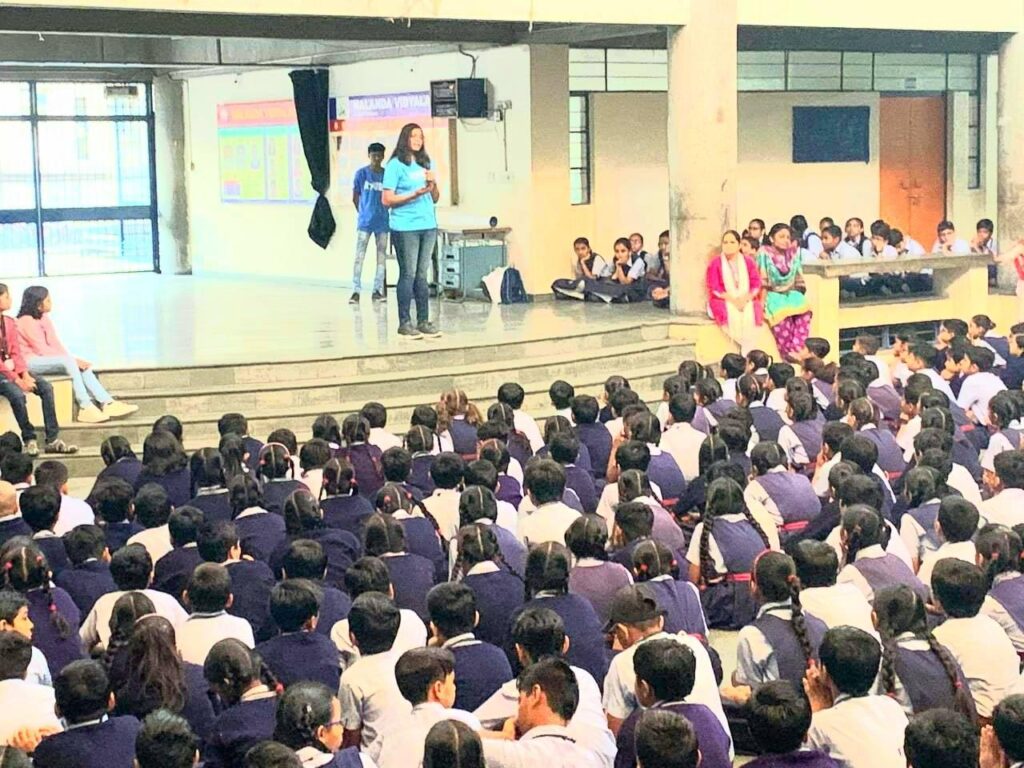 From the back of the hall, a tall AYUDH female member in jeans and a light blue AYUDH tshirt presents to the seated students.