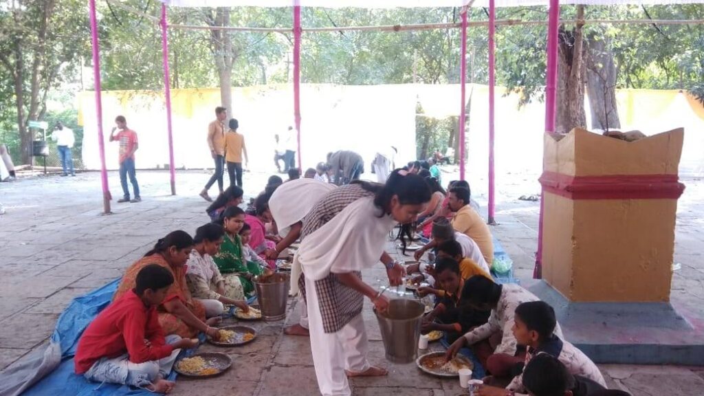 A young female AYUDH member, leans down to serve food to a young boy sitting in a row of children.