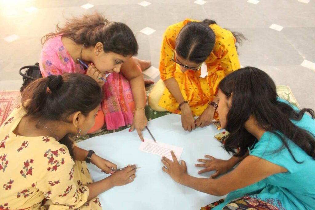 Seated on the floor, four young women dressed colorfully, design their poster board presentation on Shivaratri.