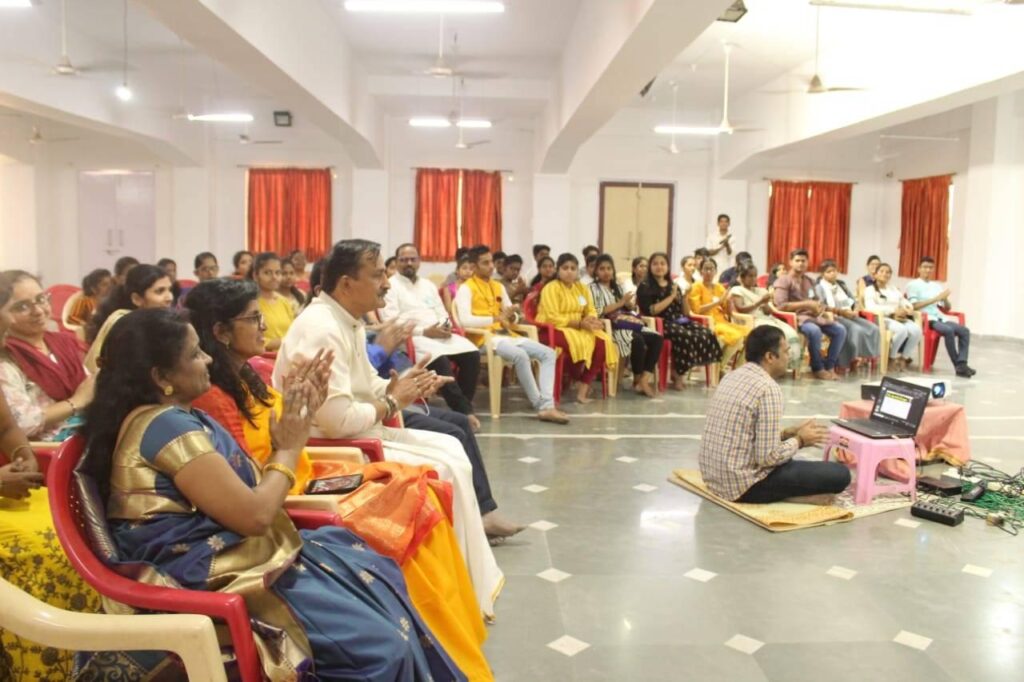 An audience of about one hundred people seated in chairs listening to the video of Shivaratri.