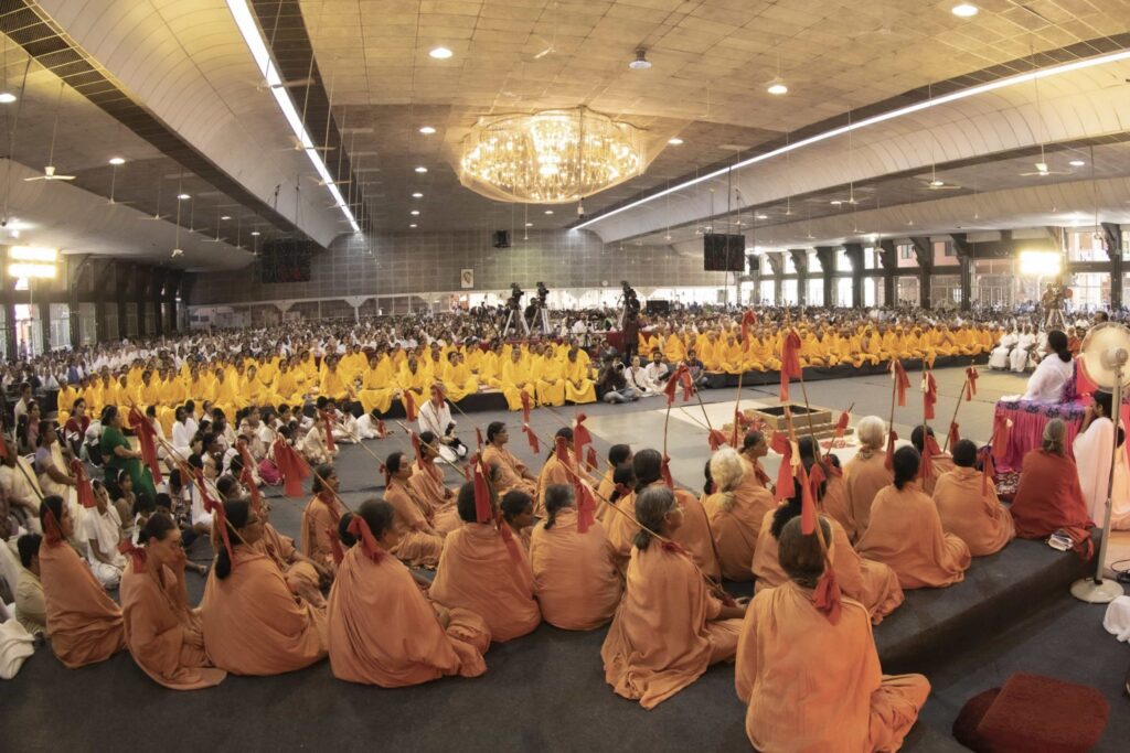 A wide view from behind, the sannyasa initiates with staff's over their right shoulders, and the several rows of the new yellow robed brahmacharya initates are seated facing Amma.