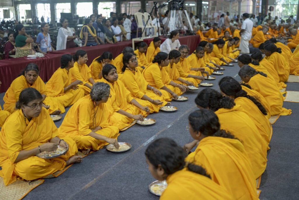 Rows of newly ordained Brahmacharinis dressed in bright yellow robes, sit facing each other eating a prasad meal.