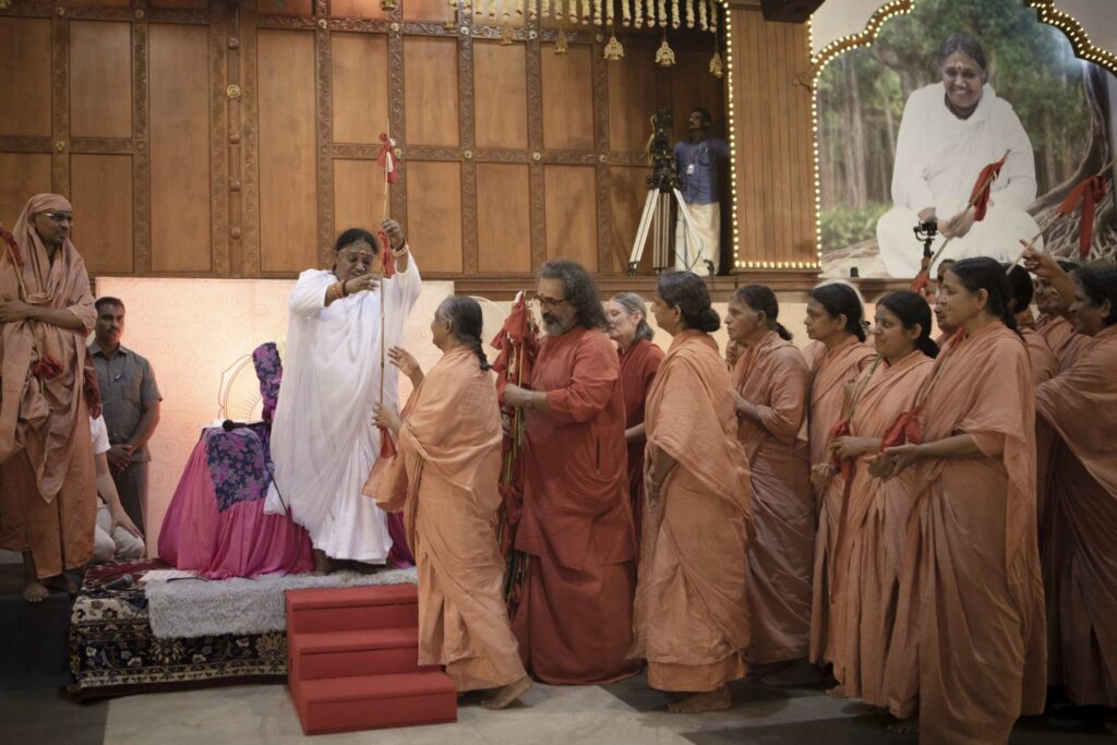 Amma is standing handing the staff with 3 flags to a female sannyasi initiate as her sisters and Swami Amritaswarupananda are next to her.