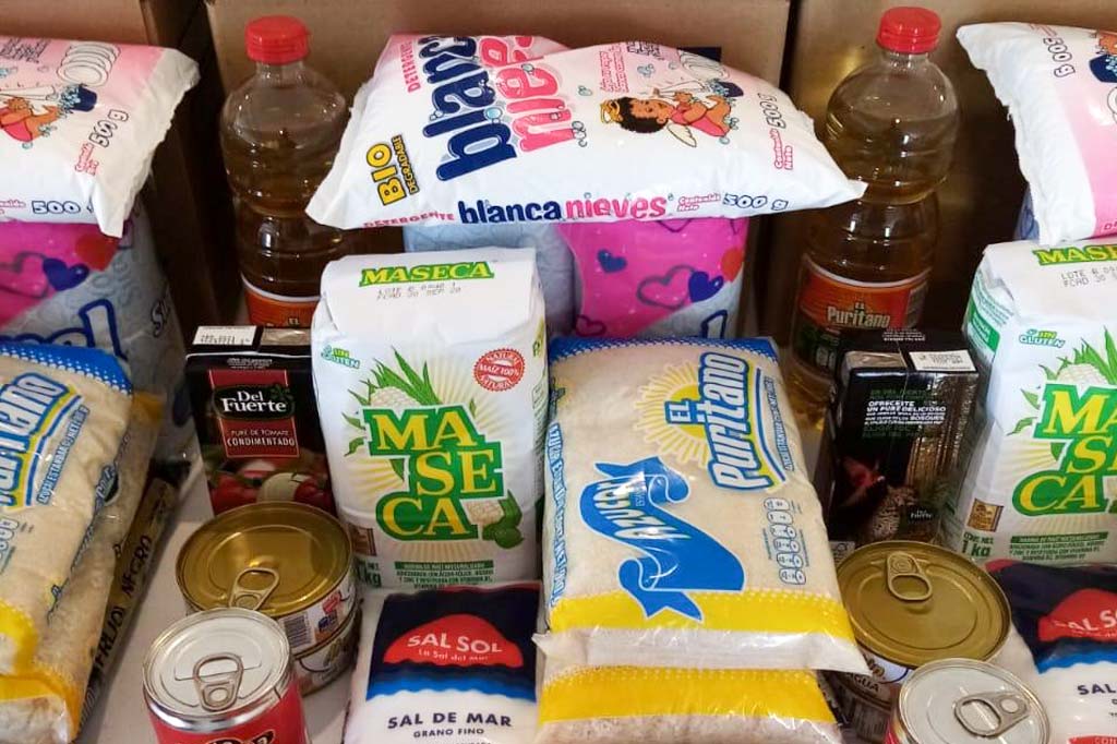 A supply box contains rice, beans, sugar, salt, oil, lentils, tuna, cookies, pasta, jalapeños, flour, toilet paper and soap.