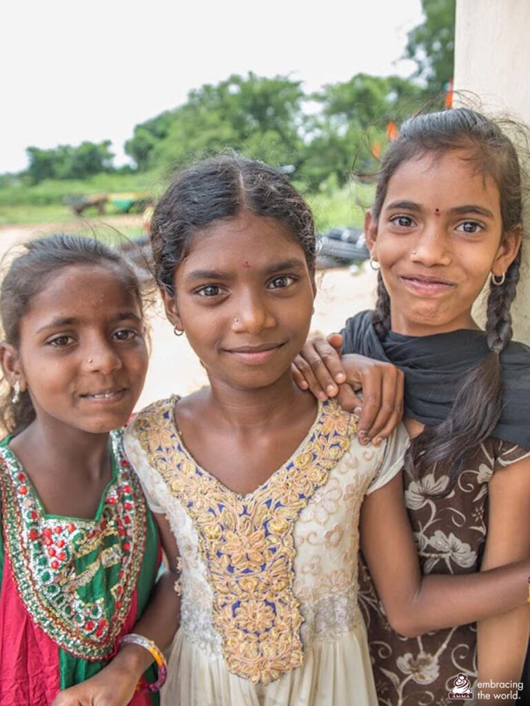 Three girls in colorful dresses stand close to each other from shortest to tallest and smile. 