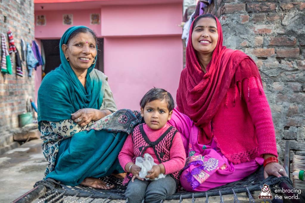 Three women sit outside on a cot- an older woman in blue, a younger woman in red, and a toddler in pink between them.