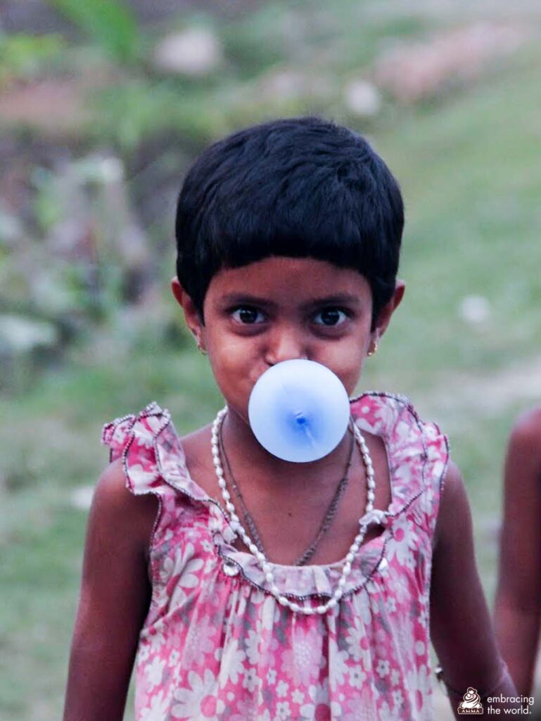 A small child in a pink dress and short hair blows a big bubble with blue bubblegum. 