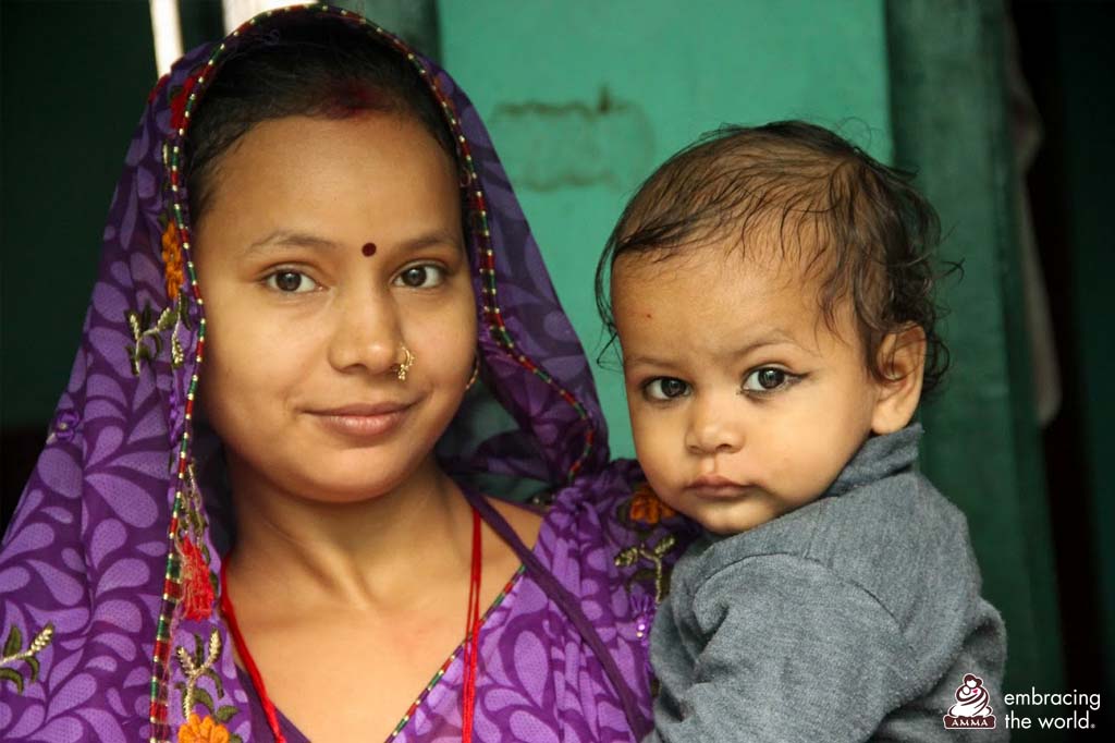 A woman in a purple sari with her head covered carries a baby with dark eyeliner in her arms.