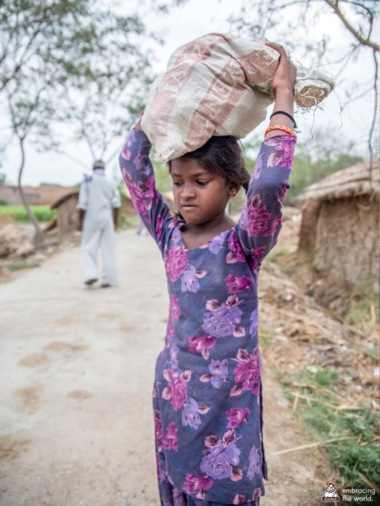 A young girl in purple looks down while walking down a road carrying a sack on her head. 