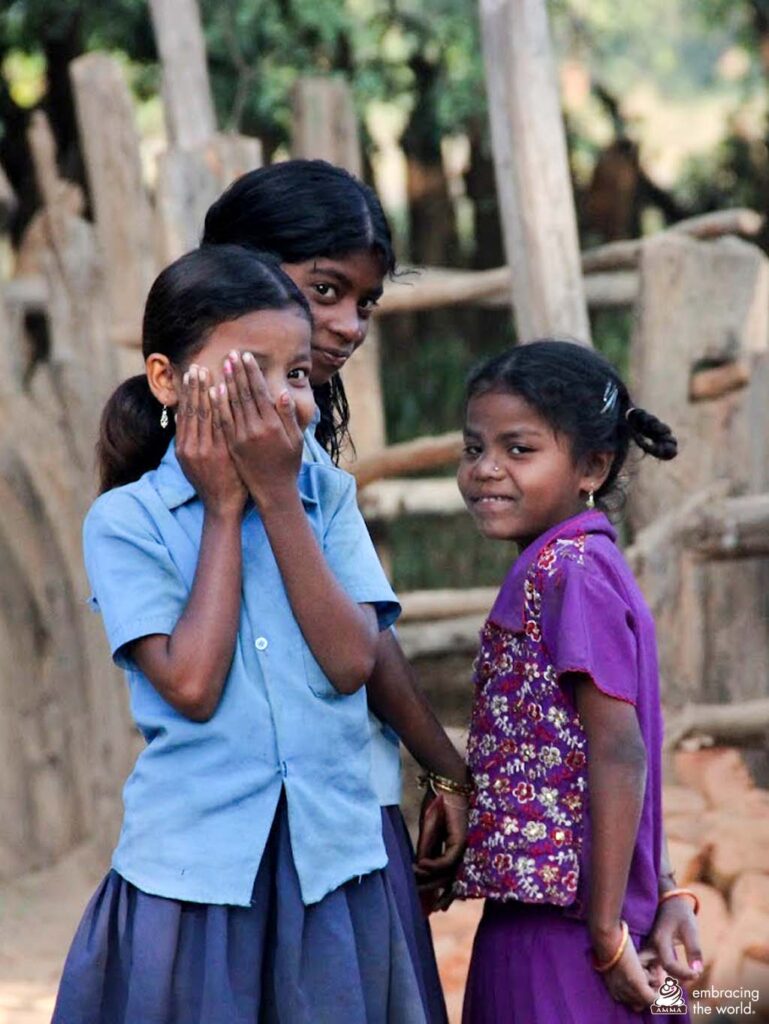 Three girls stand outside smiling. In the front, one of the girls playfully peeks out behind her hands.  