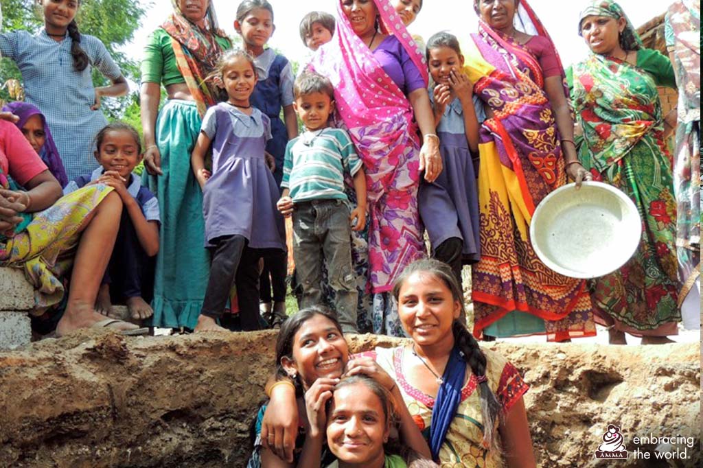 A group of villagers gather around a pit which has been dug for a toilet. Three women are in the pit, smiling