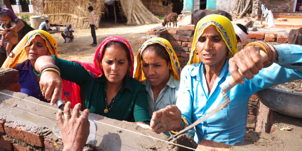 Village women with coverings on their heads for the heat, work to add cement to a brick wall.