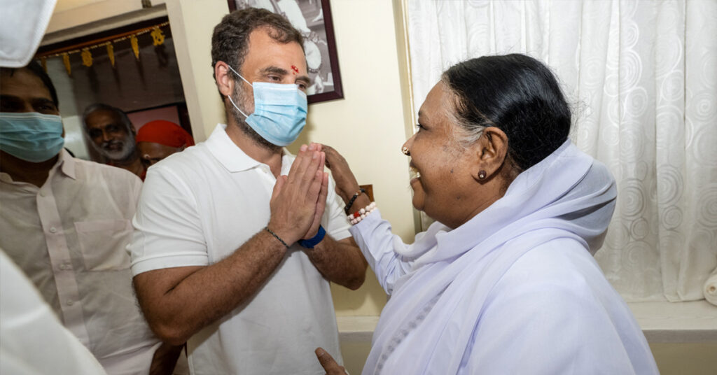 Amma and Mr. Gandhi standing. Mr. Gandhi has his palms together and Amma's hand is on his shoulder.