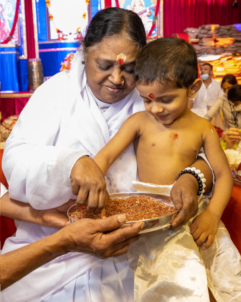 Amma sits with a child on her lap and a tray of rice, teaching him to write in the rice