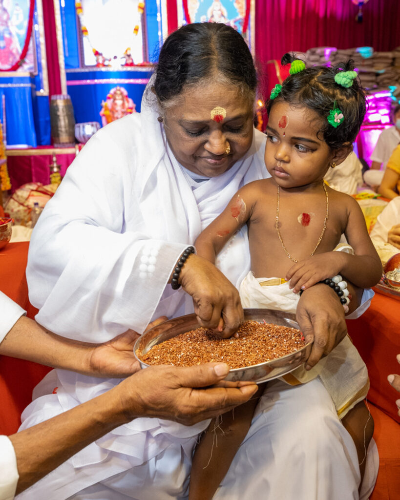 Amma teaches a baby girl to write in rice, as the girl looks at Amma's face