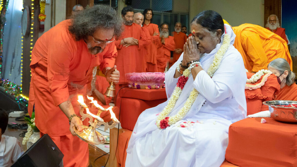 Amma, with her hands folded in prayer, surrounded by her senior disciples, as Swami Amritaswarupananda Puri waves a lamp in front of her as part of the tradition of arati.