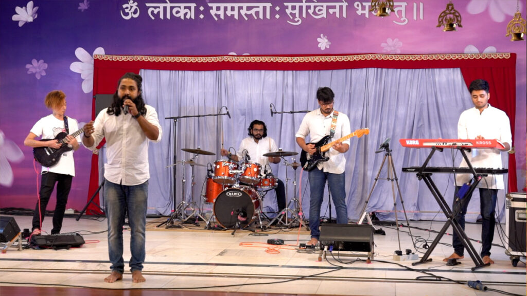  A musical performance by 5 artists - one on guitar, drums, bass guitar, keyboard, and one singer