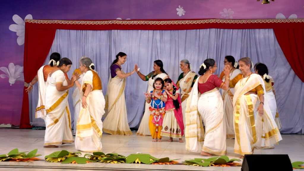 Dancers in traditional Indian outfits dance in a circle around two children dressed as Radha and Krishna 