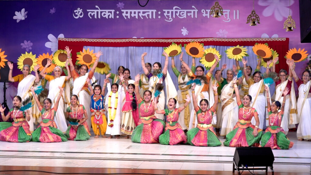 A large group of women dancers  including children on stage holding up large sunflowers 