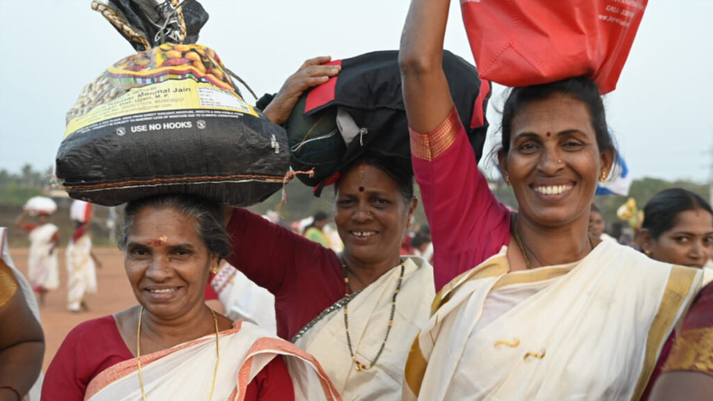 Three smiling women with sacks of supplies balanced on their heads.