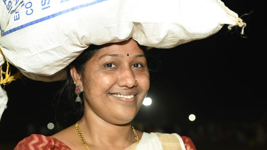 A smiling woman with a sack of supplies balanced on her head.