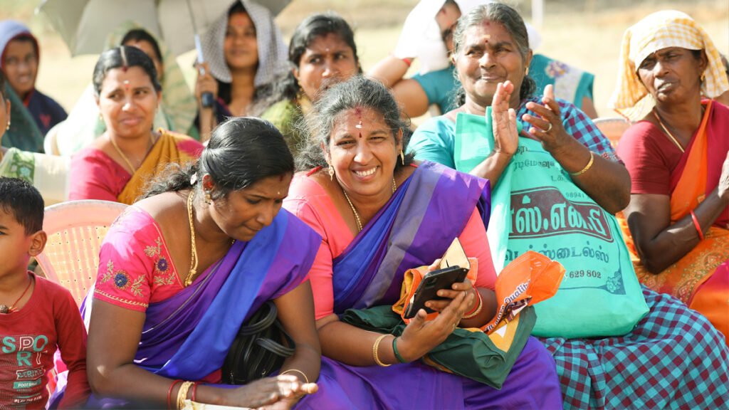 Three rows of women up close are seen in different colored saris of purple, pink, red and orange, smiling and clapping after having received their kits. 