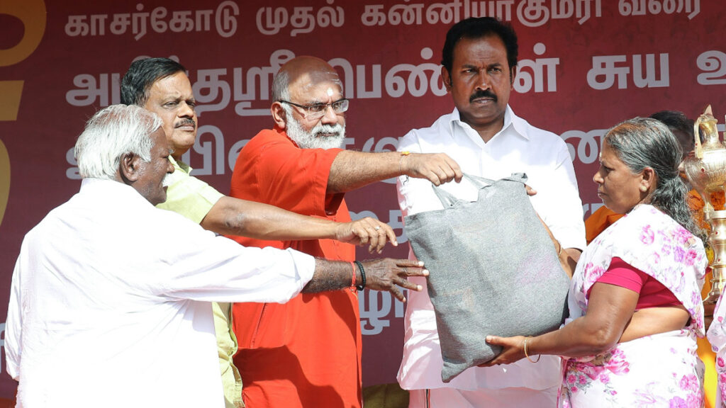 Swami Ajamritananda hands a kit to a woman in a white sari with a pink floral pattern on white. 