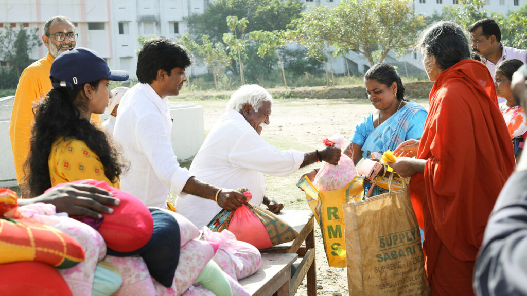 An older man dressed in white hands kit items to a woman dressed in a light blue sari with a Swamini standing next to her.