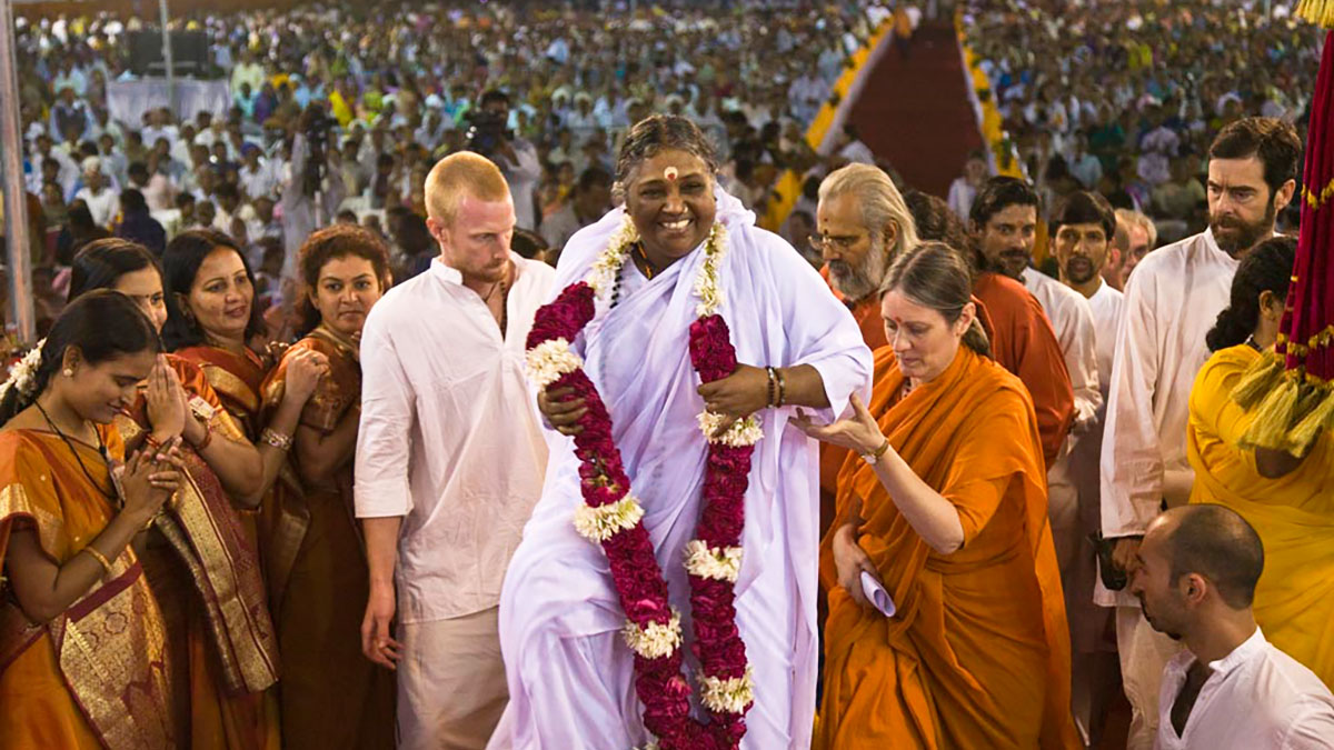 Amma arriving to the stage in a big Indian event hall filled with thousands of people, smiling radiantly, wearing a white sari and a beautiful garland of red and white flowers.