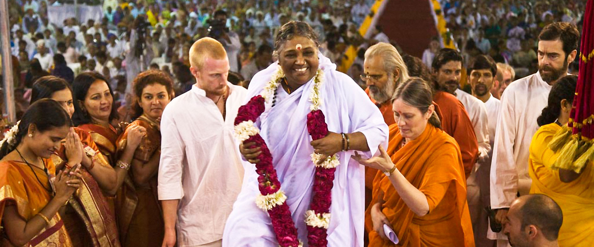 Amma arriving to the stage in a big Indian event hall filled with thousands of people, smiling radiantly, wearing a white sari and a beautiful garland of red and white flowers.