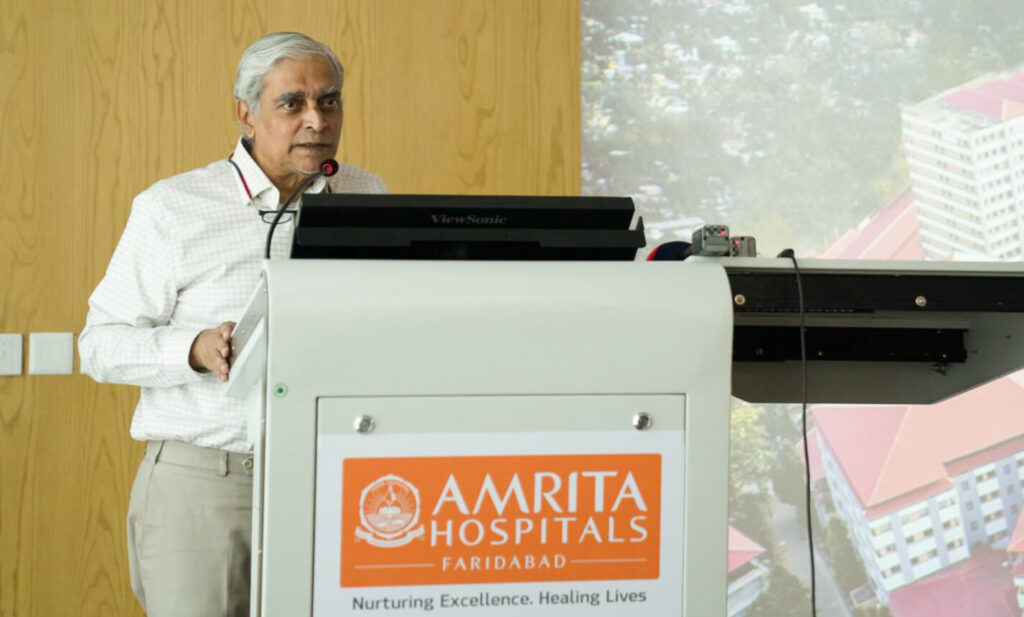 Dr. Prem Nair stands at the Amrita Hospital Faridbad podium in a white shirt and tan pants, sharing the medical care vision for the people of India.