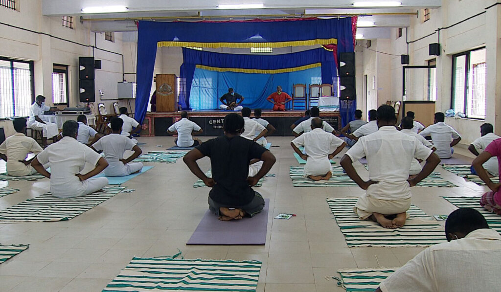 Seen from the back of hall, inmates have their hands on their waist, with knees bent, seated on heels of their feet in a yoga asana.