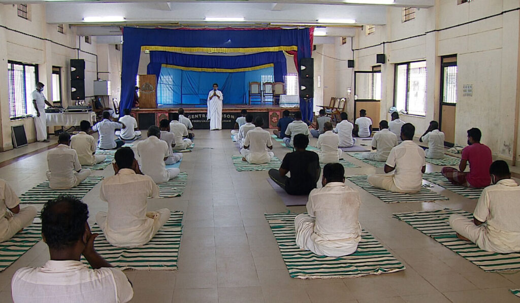 From the back of the hall, Brahmacharini Preethi instructs IAM meditation technique with inmates seated.