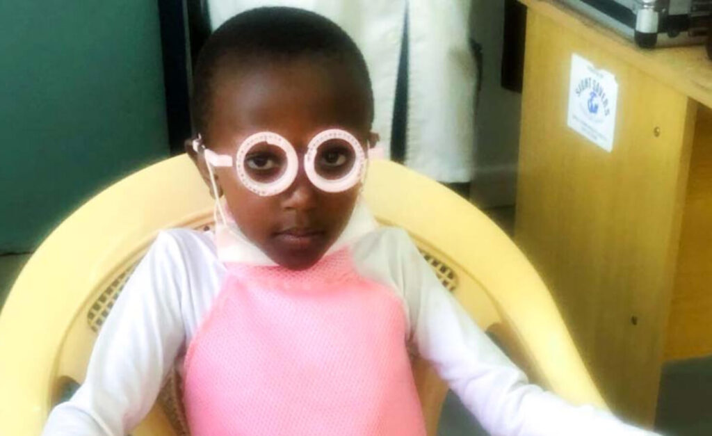 Charity sits in a yellow plastic chair, wearing a pink and white dress and round eyelasses.