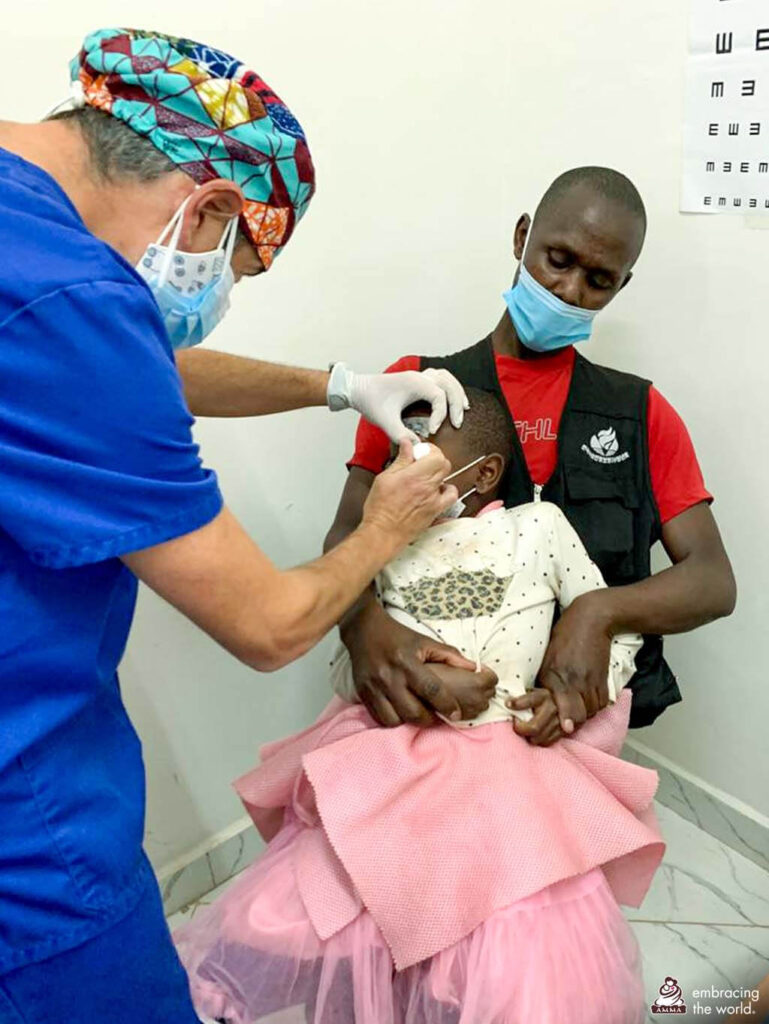 Charity in her pink and white dress, sits on her dad's lap while the doctor examines her left eye.