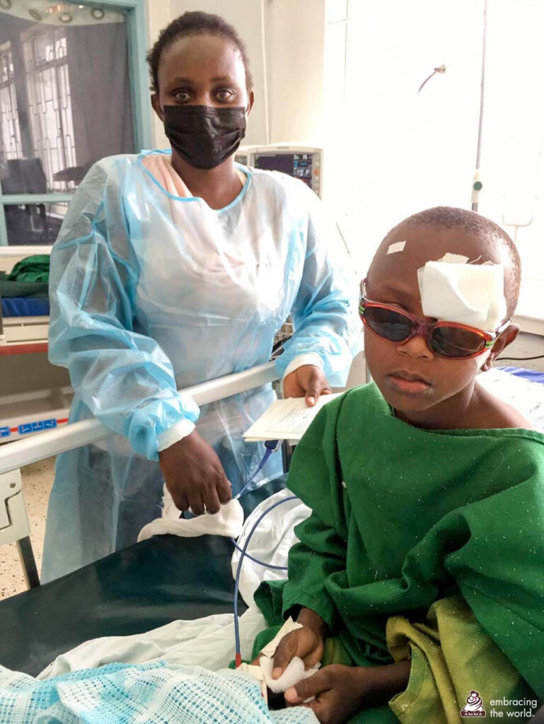 Six year old Charity sits up after surgery with a bandage over her left eye and red glasses. Her mother in a hospital gown and mask stands behind her.