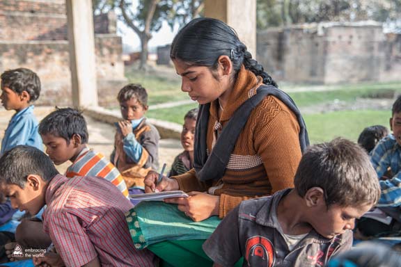 Indian woman writes in a notebook. She is seated on the floor surrounded by children