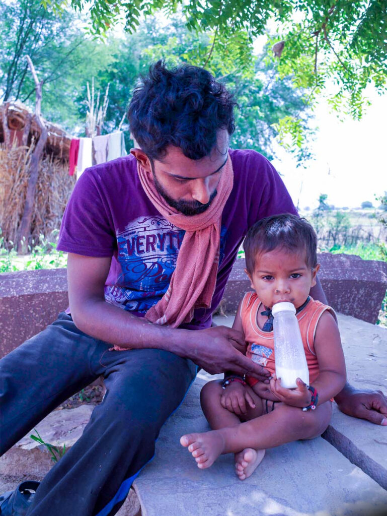 A father in a purple tshirt and mauve scarf helps feed his small child with a bottle of milk.