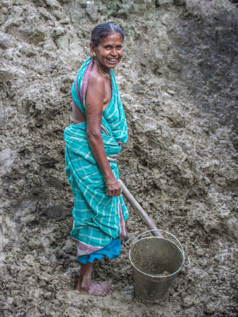 An older woman, smiling, in a blue green simple sari, stands barefoot, shoveling heavy mud into a bucket.