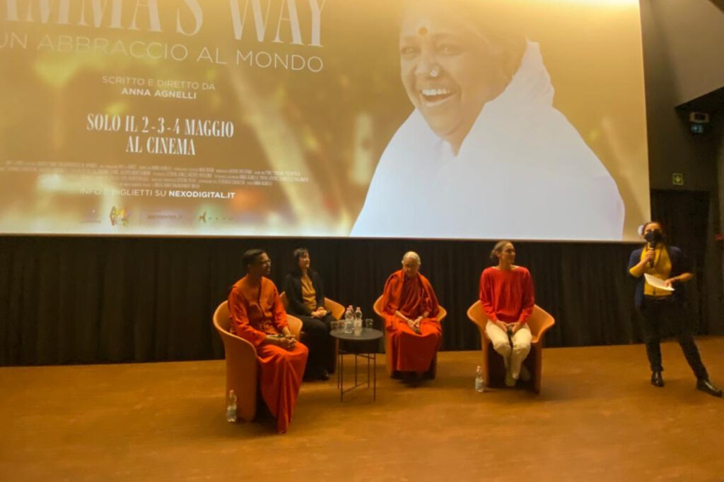 Swami Shubamritananda Puri, Manuela Ceresola with Amma Italia, Swamini Amritajyoti Prana, and filmmaker Anna Agnelli sitting on stage in front of the movie screen at the debut for the new documentary about Amma.