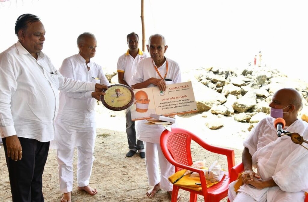 three men holding a clock and a sign representing the yatra while H.H. Acharya is seated and looking on.