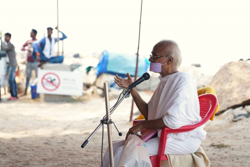 H.H. Acharya sitting in a chair talking into a microphone.